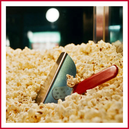 Party & Event RentalsA metal scoop with a red handle buried in a pile of freshly popped popcorn. The background is softly blurred, conveying a cozy, inviting feel.